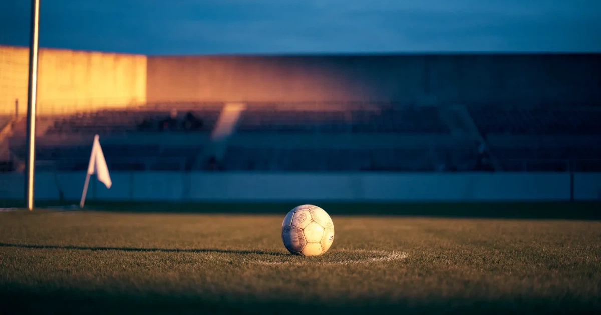 Ein Fußball liegt auf dem Anstoßpunkt eines leeren Rasenplatzes, im Hintergrund eine große Stadiontribüne im Abendlicht.