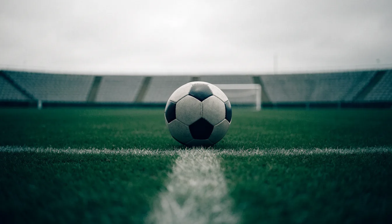 Ein Fußball liegt auf dem Anstoßpunkt eines leeren Rasenplatzes, im Hintergrund eine ruhige Tribüne unter bedecktem Himmel.