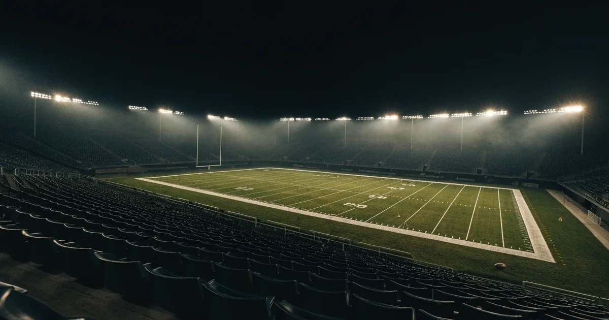 Ein American-Football-Feld mit weißen Yardlinien im Flutlicht, von einer hohen Tribüne aus gesehen, die Ränge leer, Zahlen nicht erkennbar.