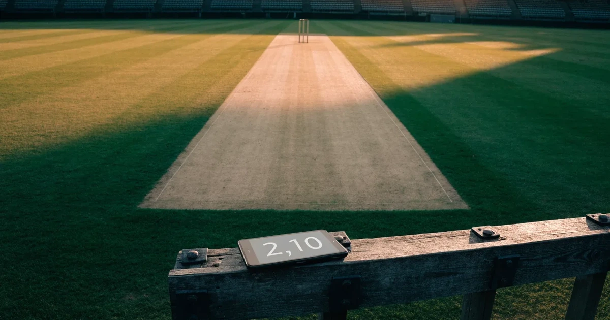 Ein weites grünes Cricketfeld mit einem hellen Streifen in der Mitte, tief stehende Sonne, im Hintergrund eine leere Tribüne.