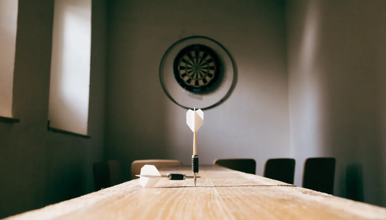 Eine Wurflinie aus Holz auf dem Hallenboden, im Hintergrund in der Ferne eine Dartscheibe an der Wand.