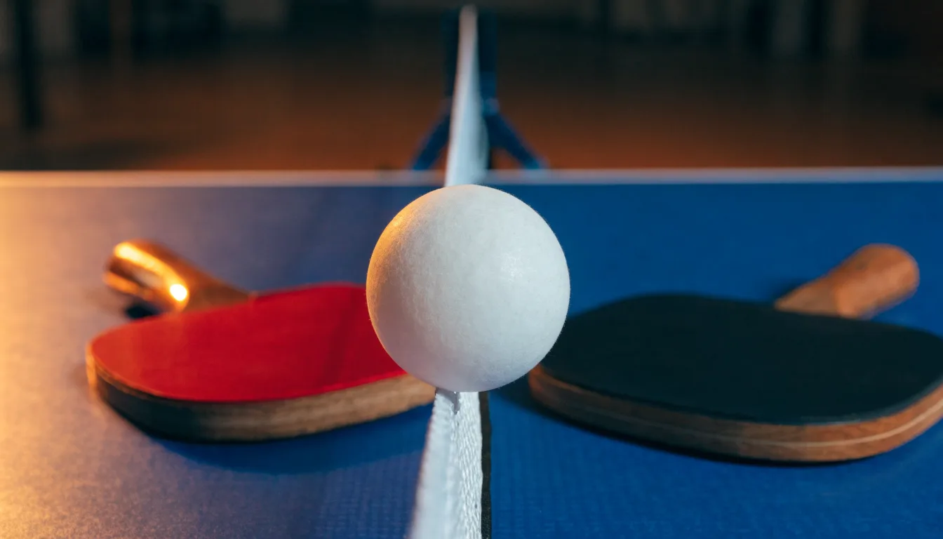 Ein weißer Ball liegt in Nahaufnahme auf der Netzkante einer Tischtennisplatte, daneben zwei Holzschläger mit rotem und schwarzem Belag.