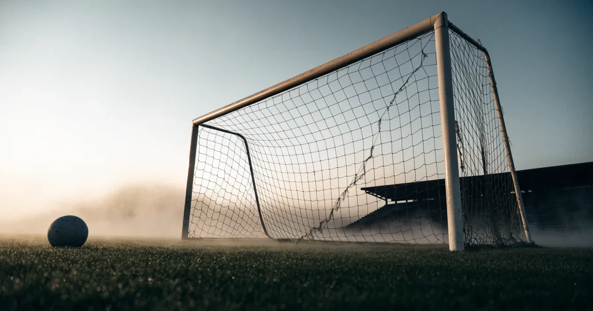 Ein Fußballtor mit Netz steht im Morgennebel auf einem leeren Rasenplatz, im Hintergrund die Silhouette einer kleinen Tribüne.