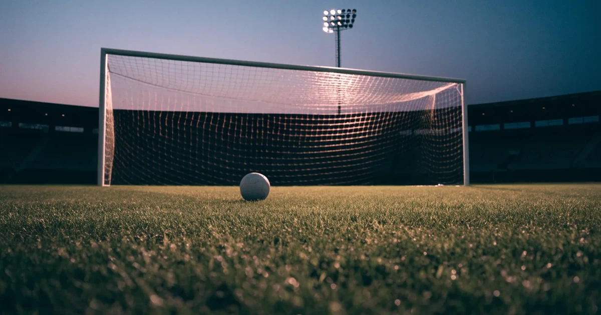 Ein Fußballtor mit Netz im Gegenlicht auf einem leeren Platz am Abend, davor ein Ball auf dem Rasen.