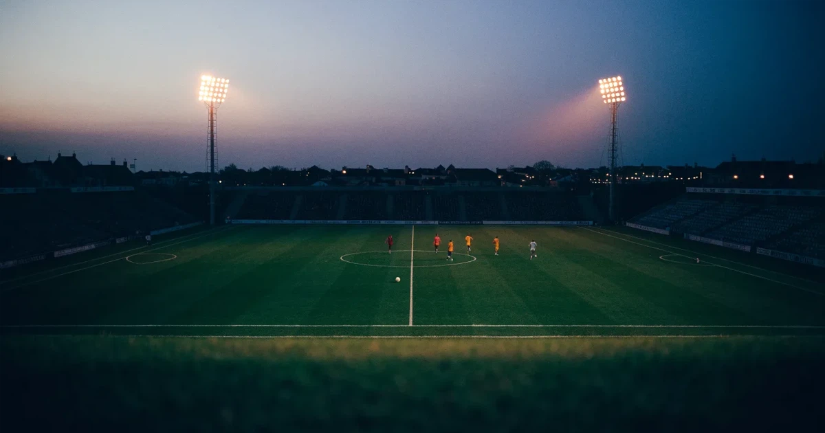 Ein Fußballstadion in der Dämmerung aus der Ferne, die Flutlichter gerade eingeschaltet, davor ein Kreisverkehr mit Laternen.