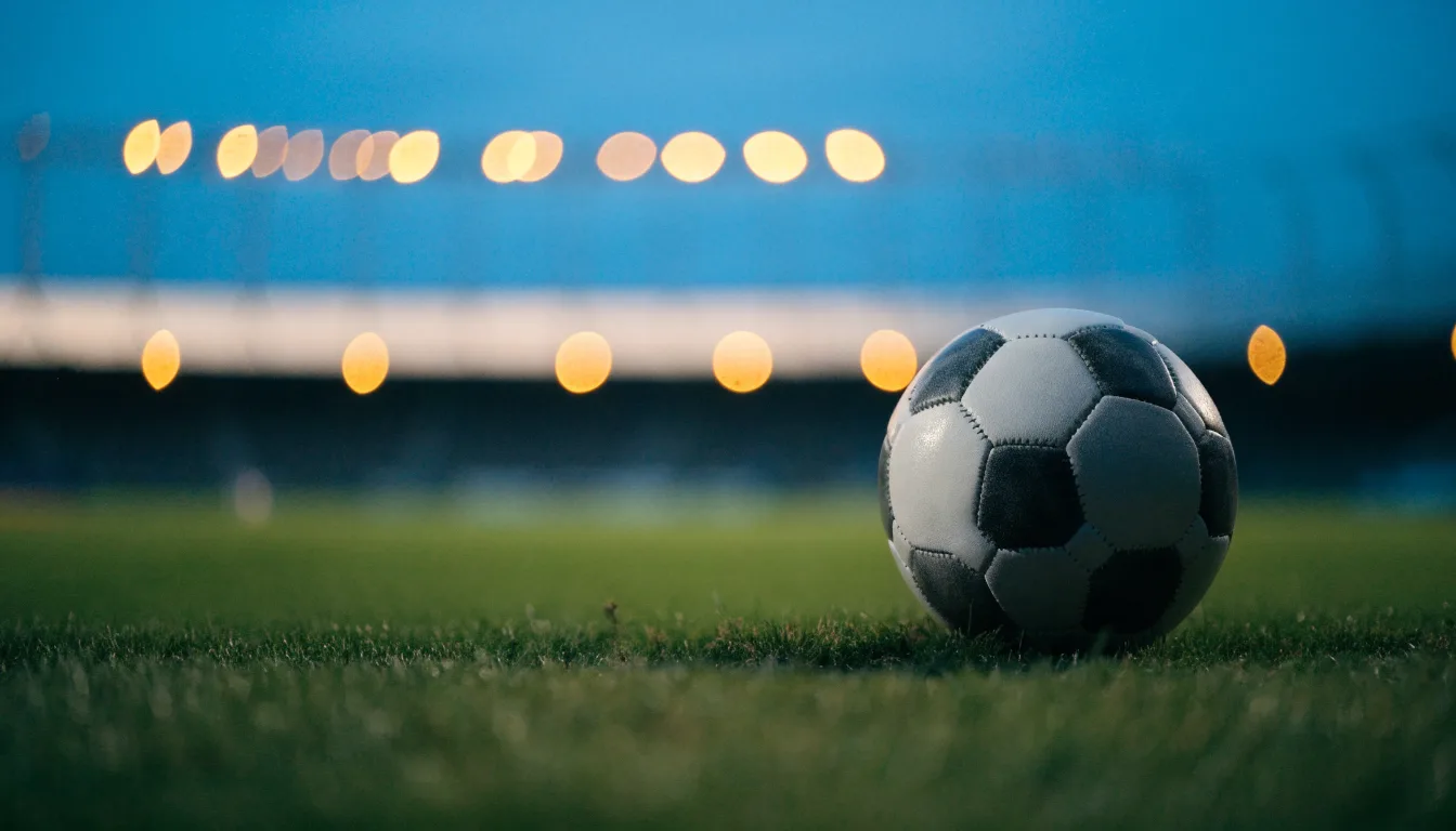 Ein Fußball liegt auf einem Rasenplatz in der Dämmerung, im Hintergrund ein ferner Stadionrand unter Flutlicht.
